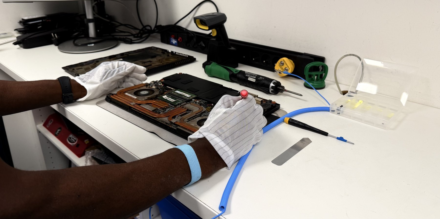 Les mains d’un technicien, en gants et bracelet antistatiques, sur un ordinateur portable ouvert : capot déposé, ventilateurs, caloducs et barrette mémoire apparents.