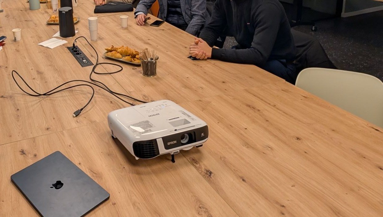 Une table de réunion en bois clair : un vidéoprojecteur relié par un câble à une prise encastrée, un plateau de viennoiseries, des gobelets et un ordinateur portable fermé.